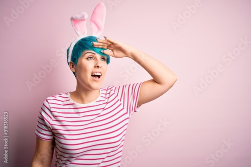 Young woman with fashion blue hair wearing easter rabbit ears over pink background very happy and smiling looking far away with hand over head. Searching concept.