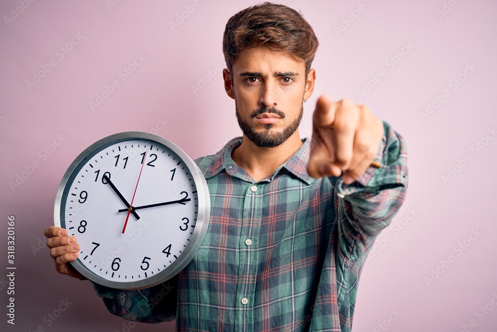 Young man with beard doing countdown using big clock over isolated pink background pointing with finger to the camera and to you, hand sign, positive and confident gesture from the front Young man with beard doing countdown using big clock over isolated pink background pointing with finger to the camera and to you, hand sign, positive and confident gesture from the front