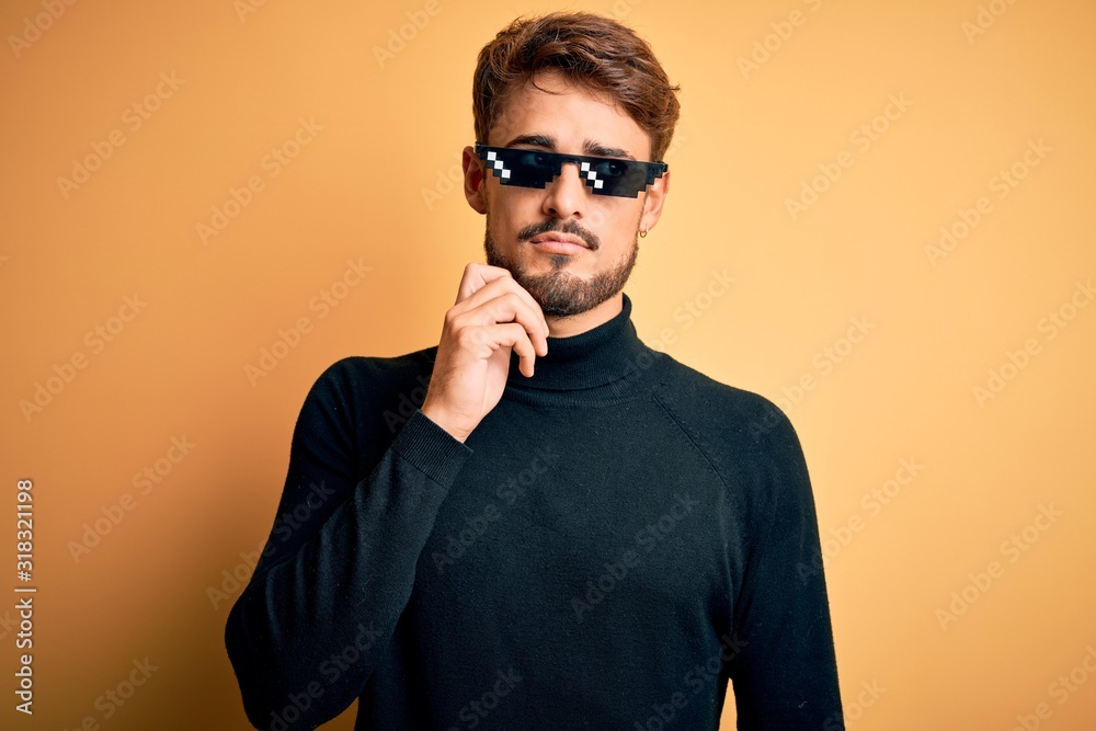 Obraz premium Young man wearing thug life fanny sunglasses standing over isolated yellow background with hand on chin thinking about question, pensive expression. Smiling with thoughtful face. Doubt concept.