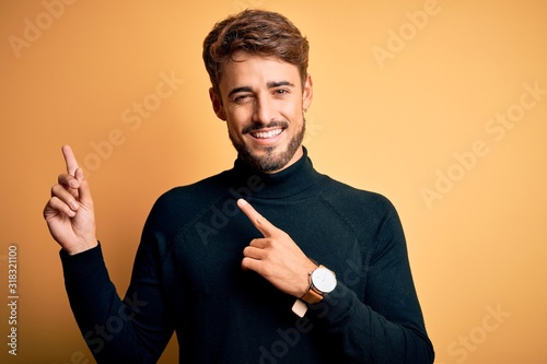 Young handsome man with beard wearing turtleneck sweater standing over yellow background smiling and looking at the camera pointing with two hands and fingers to the side.