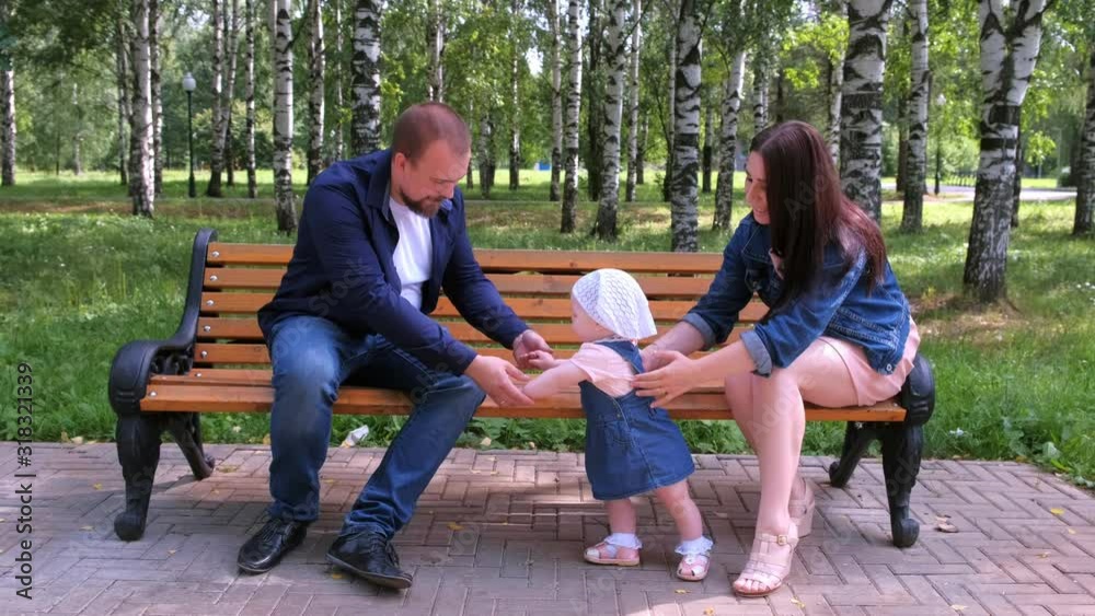 Happy family walking at weekend. Parenthood and baby care. Baby girl is trying to make first steps from mom to dad holding hands sitting on bench in park. Parents learning daughter to walk.