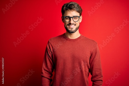 Young handsome man with beard wearing glasses and sweater standing over red background with a happy and cool smile on face. Lucky person.