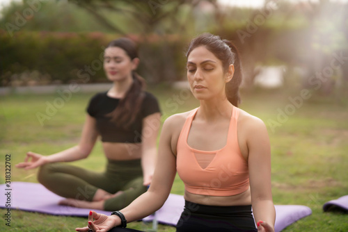 Two beautiful brunette woman wearing tight active wear performing yoga poses in a park on purple mats with soft sun flares coming through the trees 