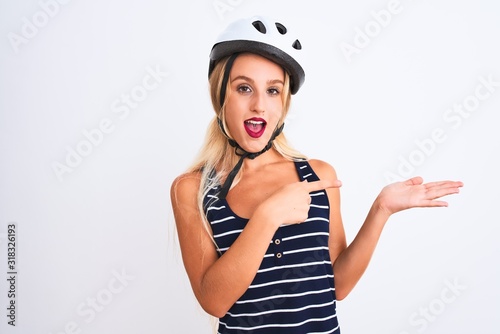 Young beautiful woman wearing bike helmet standing over isolated white background amazed and smiling to the camera while presenting with hand and pointing with finger. © Krakenimages.com
