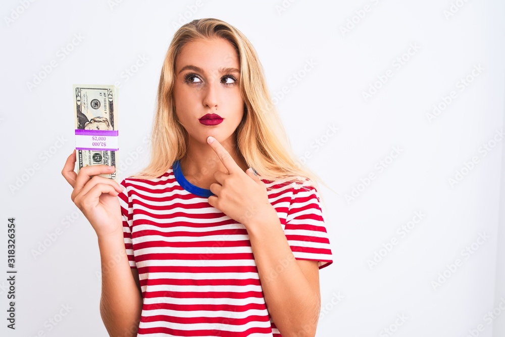 Beautiful woman wearing red striped t-shirt holding dollars over isolated white background serious face thinking about question, very confused idea Beautiful woman wearing red striped t-shirt holding dollars over isolated white background serious face thinking about question, very confused idea
