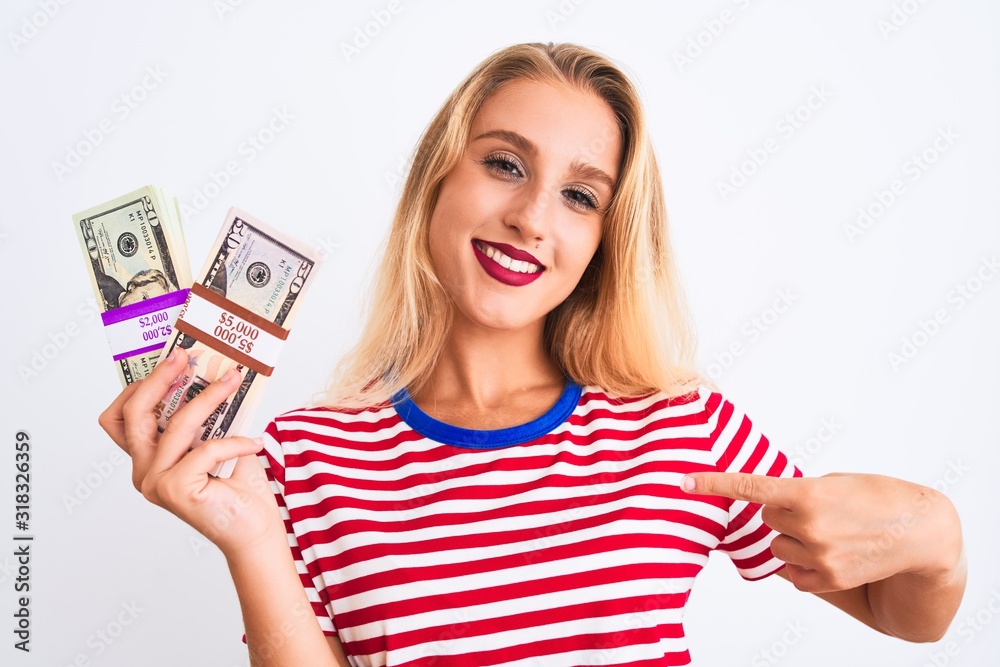 Beautiful woman wearing red striped t-shirt holding dollars over isolated white background with surprise face pointing finger to himself Beautiful woman wearing red striped t-shirt holding dollars over isolated white background with surprise face pointing finger to himself