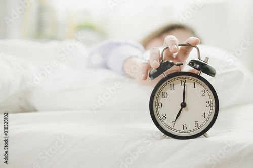 young asian woman sleeping on bed pressing snooze button on black vintage alarm clock at seven o'clock morning in bed room at home, lifestyle, good morning, healthy sleep and joyful weekend concept