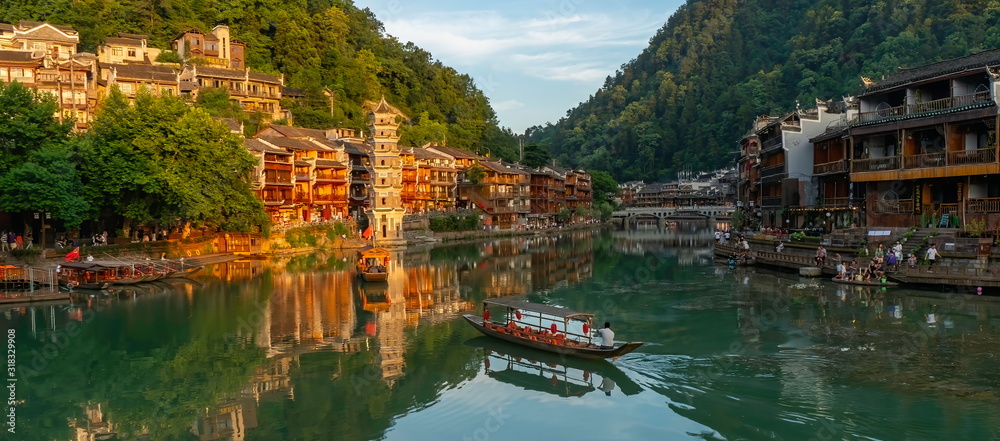 Picturesque panorama of an authentic Chinese landscape. Traditional boat sails the waters of the Tuojiang River with the old Wanmingta Pagoda, old wooden houses. Fenghuang Ancient Town, China