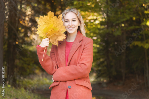 Portrait of attractive caucasian young blonde girl in red coat with a bouquet of fallen yellow leaves smiling in the autumn forest. The concept of autumn and fall holidays and weekends