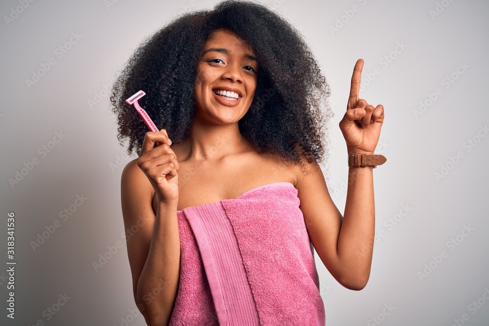 Young african american woman with afro hair in a towel holding female razor for body depilation surprised with an idea or question pointing finger with happy face, number one Young african american woman with afro hair in a towel holding female razor for body depilation surprised with an idea or question pointing finger with happy face, number one