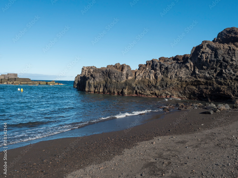 The dark sand beach of the village of Los Cancajos at La Palma, Canary Islands. Lava rock cliffs and Blue sky background.