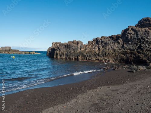 The dark sand beach of the village of Los Cancajos at La Palma, Canary Islands. Lava rock cliffs and Blue sky background.