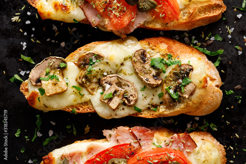 Grilled bruschetta with the addition of tomatoes, capers, ham, mushrooms and herbs on a black background, top view , close up