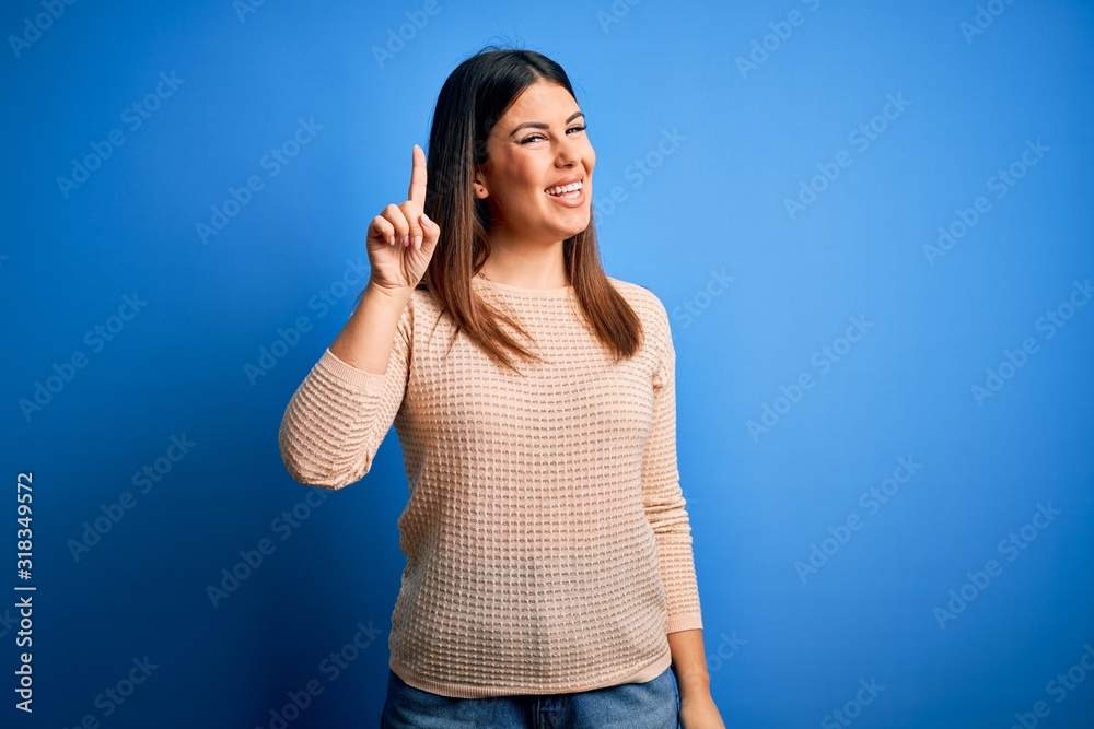 Fototapeta premium Young beautiful woman wearing casual sweater over blue background pointing finger up with successful idea. Exited and happy. Number one.