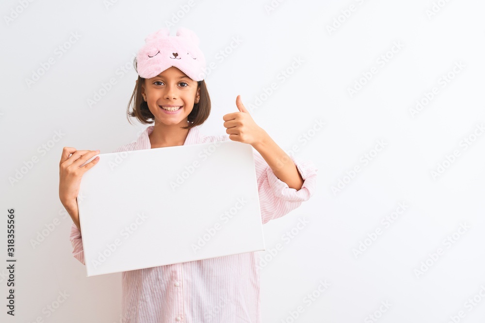Child girl wearing sleep mask and pajama holding banner over isolated white background happy with big smile doing ok sign, thumb up with fingers, excellent sign Child girl wearing sleep mask and pajama holding banner over isolated white background happy with big smile doing ok sign, thumb up with fingers, excellent sign