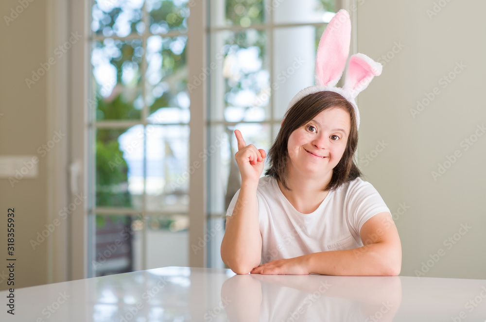 Down syndrome woman at home wearing easter rabbit ears surprised with an idea or question pointing finger with happy face, number one