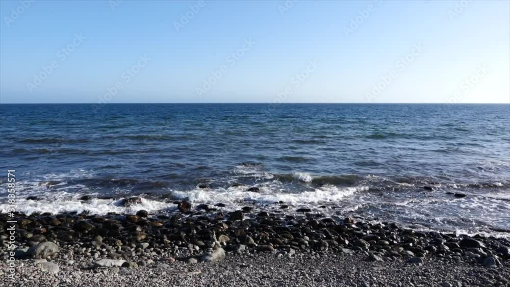 A still shot of the waves slowly crashing on the pebble shore on a beach in summer with clear blue skies A still shot of the waves slowly crashing on the pebble shore on a beach in summer with clear blue skies