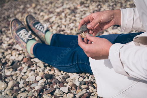 Wallpaper Mural Woman sitting on a rocky beach wearing colorful shoes, blue jeans and white raincoat, holding rocks in her hands, relaxation Torontodigital.ca