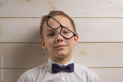 A fair-haired boy of 10 years in a white shirt with the self-tie bow tie with a serious expression on the background of a wooden wall. Black glasses without lenses.
