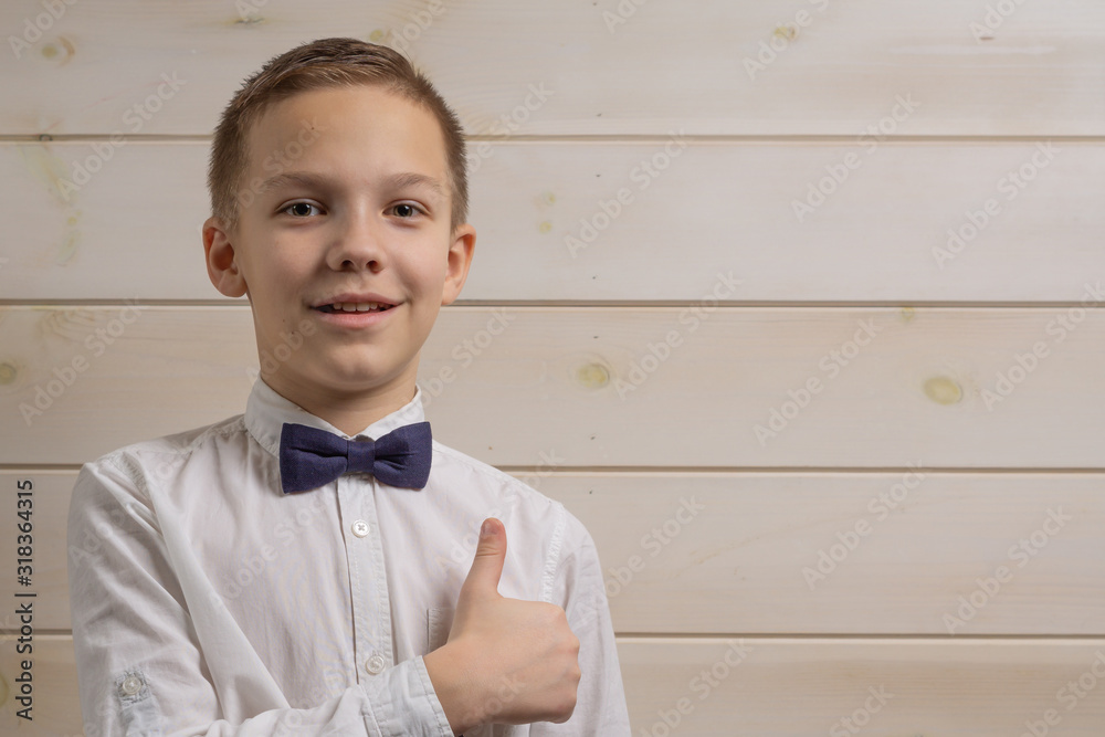 A fair-haired boy of 10 years in a white shirt with the self-tie bow tie smiles against the background of a wooden wall. Like.