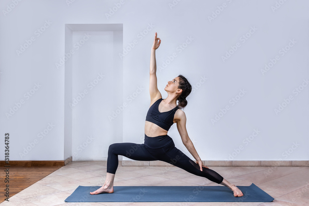 Side view portrait of brunette young woman wearing sport clothes working out against white wall, doing yoga or pilates exercise. Standing in Warrior pose, Virabhadrasana. Full length, copy space