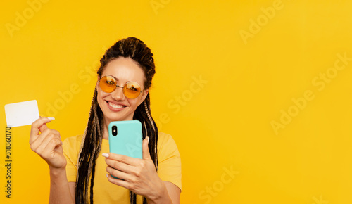 Pretty young lady in bright clothes watching camera and posing with blue phone and credit card