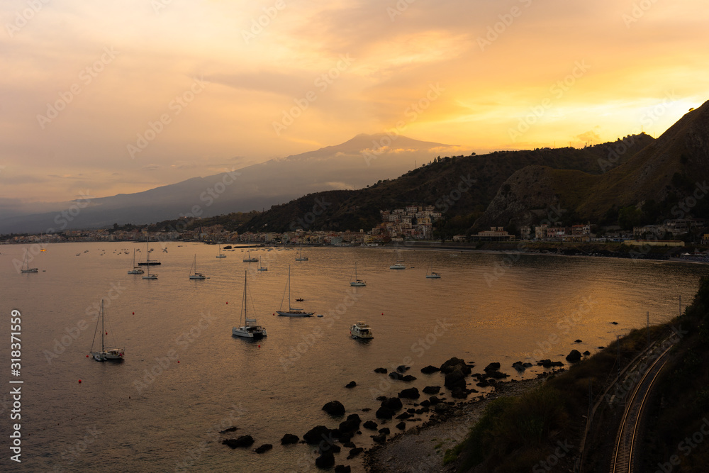 view of Mount Etna volcano with a town, port and boats below it during sunset (Sicily, Italy) 