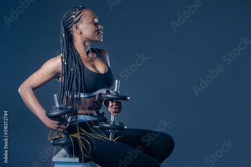 African-American model with dumbbells on a dark background. Girl with braids involved in sports. Black athlete in a fitness club. Sale of goods for sports. Sports equipment. Healthy lifestyle.