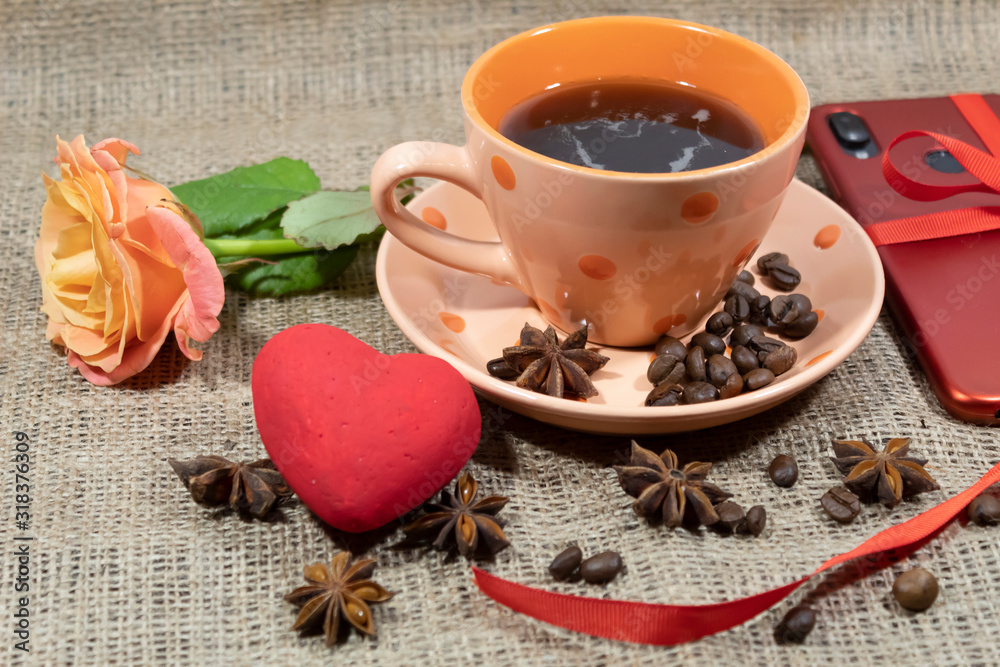 On the table is a cup of coffee. Nearby lies a rose, a red heart and a smartphone is visible. On a saucer and on a table are coffee beans and star anise. On the table is a cup of coffee. Nearby lies a rose, a red heart and a smartphone is visible. On a saucer and on a table are coffee beans and star anise.