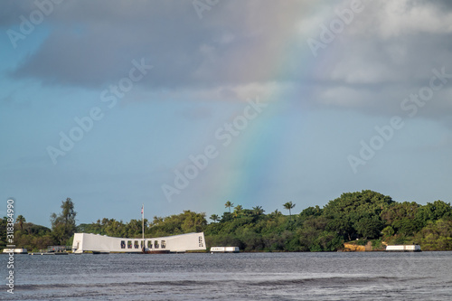 Oahu, Hawaii, USA. - January 10, 2020: Pearl Harbor. White USS Arizona Memorial with rainbow landing on it. Green belt of foliage in back separating gray sea and light blue cloudscape.