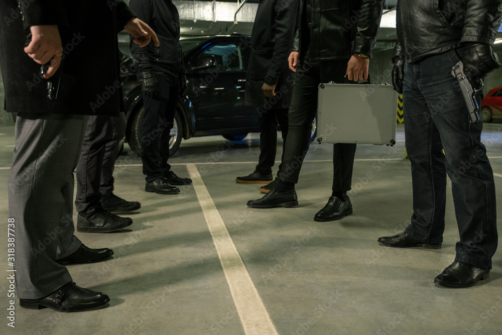 Two rows of men in black with handguns and suitcase standing on parking area