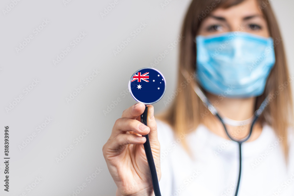 Φωτογραφία Young woman in a medical mask holds a stethoscope with the flag of Australia on a light background