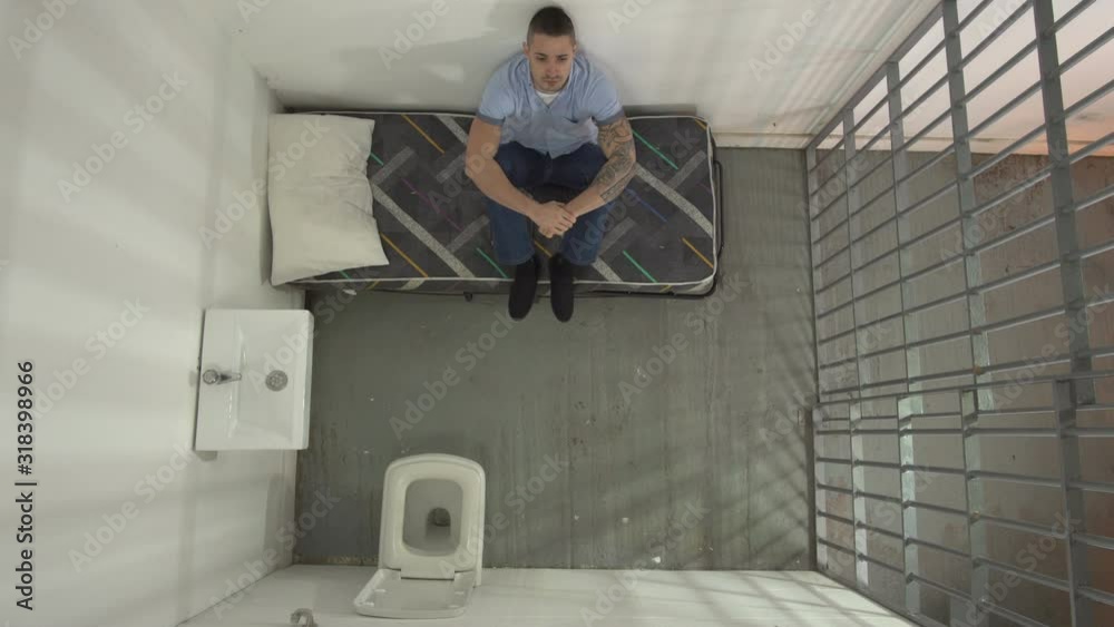Male Prisoner in Prison Cell. The inmate is sat on his bed. Aerial view from above Male Prisoner in Prison Cell. The inmate is sat on his bed. Aerial view from above