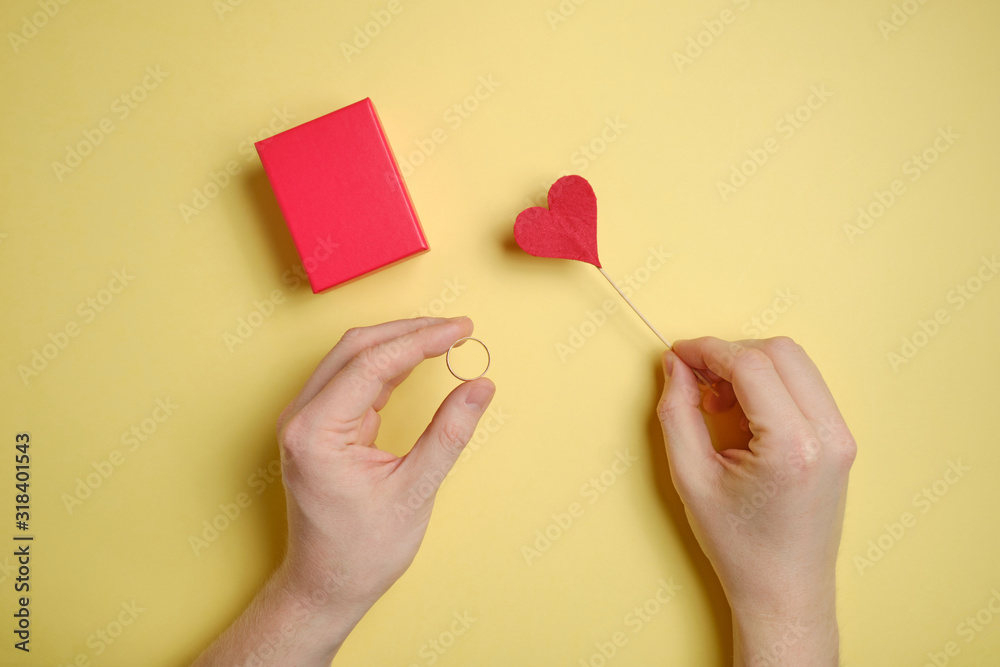 Hand holds a gold ring near a red gift box with a heart on a yellow paper background.