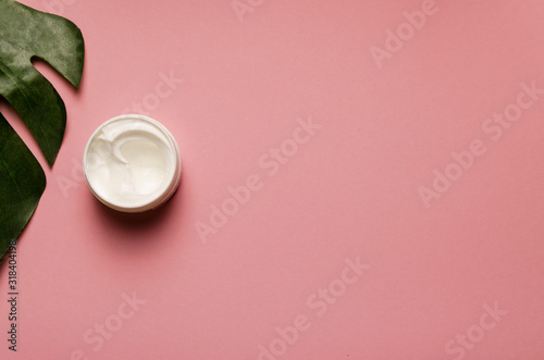 The Bank's with a white cream,the cover is open,on a pink background,cosmetology, green leaf.beauty and skin care.Top view