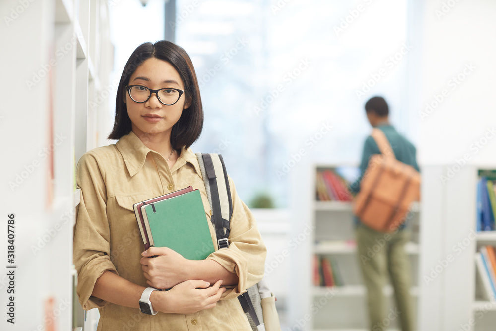 Waist up portrait of female Asian student wearing glasses and looking at camera while posing by shelves in college library, copy space Waist up portrait of female Asian student wearing glasses and looking at camera while posing by shelves in college library, copy space