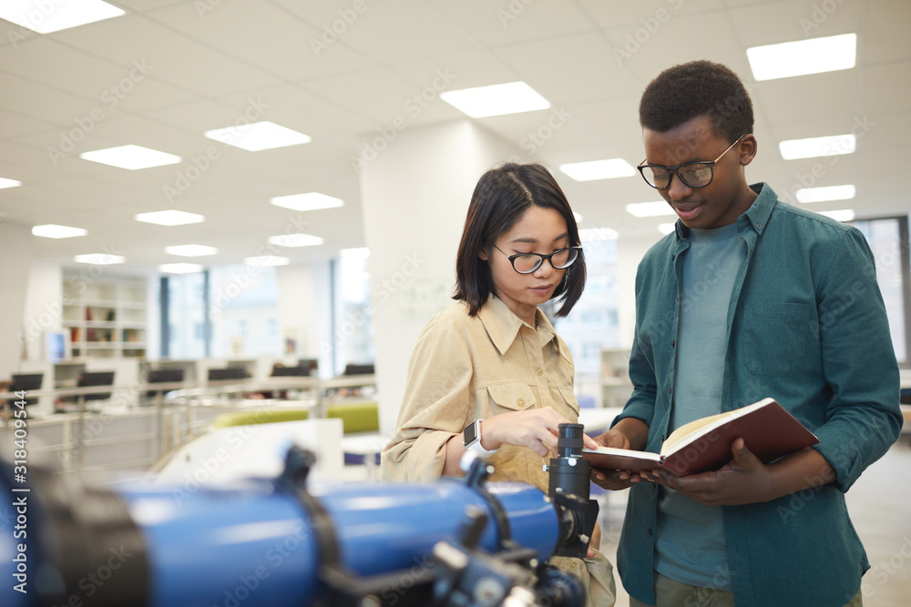 Waist up portrait of two international students reading book while standing by telescope in class or college library, copy space Waist up portrait of two international students reading book while standing by telescope in class or college library, copy space