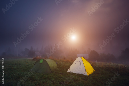 A mysterious view of the foggy countryside in the morning. Sunlight passes through the thick fog. Tents on the foggy field. Pine woods covered with fog.