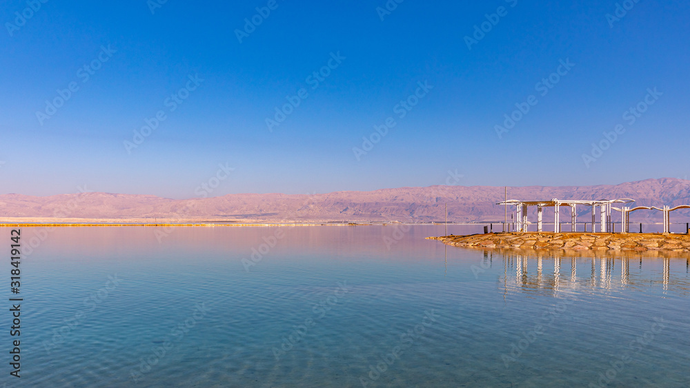 Scenic view at calm waters of  Dead Sea, Israel.  Sea beach with beach structure reflecting in calm water against blue sky and mountains in the background