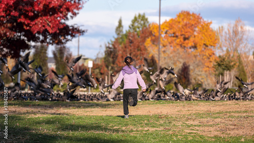 фотографія Little girl in a pick jacket and black jeans chasing geese sitting on a field in