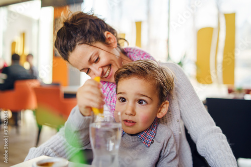 Portrait of small three years caucasian boy kid child sitting by the table with his mother young woman female at cafe having a glass of water draining lemon making lemonade