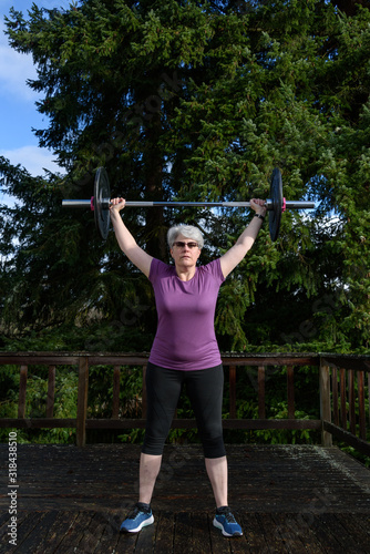 Outdoor fitness, gray haired, middle aged, woman exercising with barbell outside on deck with evergreen trees in background