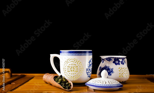 Chinese tea ceremony. Ceramic tea cup and jug with green oolong tea Tieguanyin and scoop on a bamboo desk, black background. 