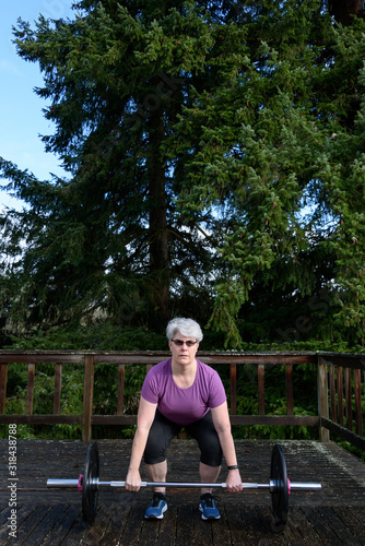 Outdoor fitness, gray haired, middle aged, woman exercising with barbell outside on deck with evergreen trees in background