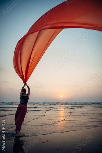 Elegant young girl performs dance moves on the sand holding a long red cloth against the background of evening sky and sun setting over the sea. Silhouette of slim woman against the sunset.
