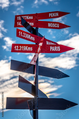 Red crossroad signpost showing the distance of several places on earth with a partially cloudy blue sky in the background. Pekin, Moscow, Paris, Madrid. © Andres Conema
