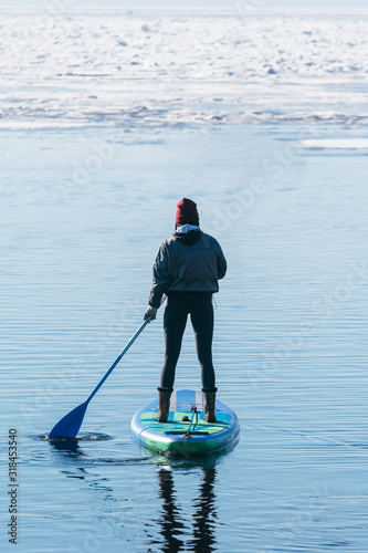 Silhouette of unrecognizable woman in wetsuit paddling on a SUP board. Female floating on a stand up paddle board in the gulf. Winter season and active leisure concept. Dynamic recreation. 