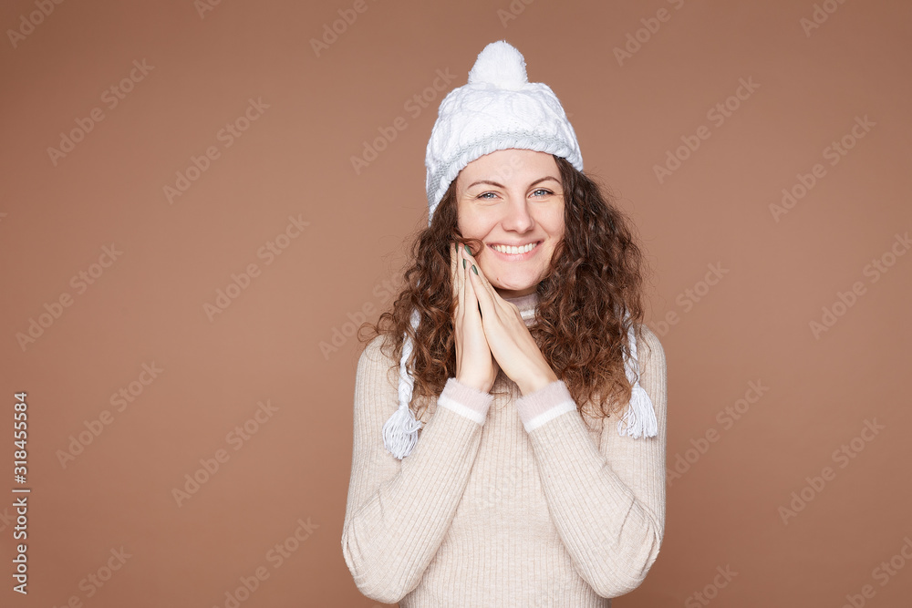 Pretty happy European woman dressed in cozy outfit, keeps hands on cheeks, wears white knitted hat, cashmere sweater, poses on beige studio wall. Cheerful lovely feminine lady feels amused, isolated. Pretty happy European woman dressed in cozy outfit, keeps hands on cheeks, wears white knitted hat, cashmere sweater, poses on beige studio wall. Cheerful lovely feminine lady feels amused, isolated.