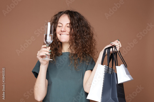 Young excited female with festive hairstyle, feels relaxed, rising glass with cold non alcoholic drink or water, looks happily, holding shopping bags. People, lifestyle, leisure and  holidays concept. photo