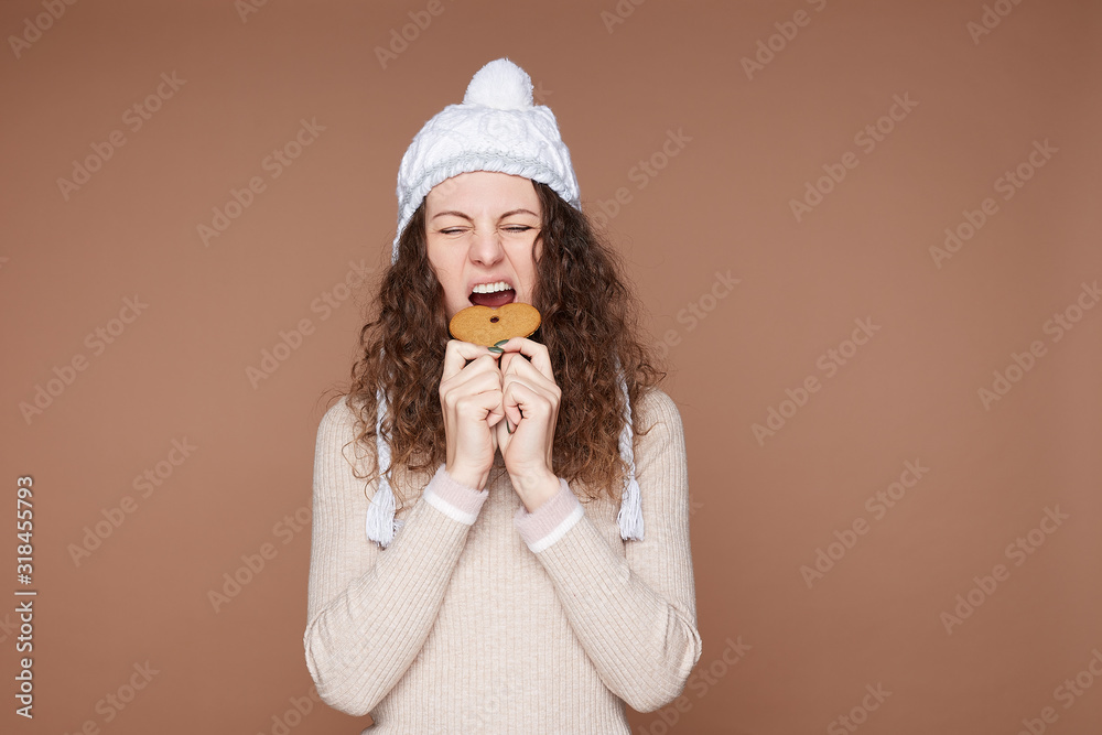 Pretty young female with curly hair holds tasty sweet ginger cookie, containing much sugar, grimacing pretending not like it, dressed in casual cashmere sweater and knitted hat. Junk food concept. Pretty young female with curly hair holds tasty sweet ginger cookie, containing much sugar, grimacing pretending not like it, dressed in casual cashmere sweater and knitted hat. Junk food concept.